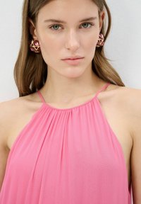 Top halter de color rosa con pliegues y escote fruncido, con tirantes delgados. La modelo lleva grandes pendientes de oro y joyas rosas, mostrando colores vibrantes.