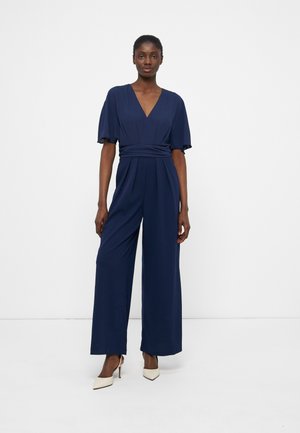 Kvinde står iført en marineblå jumpsuit med vide ben, korte flagrende ærmer og hvide spidse høje hæle mod en ensfarvet hvid baggrund.