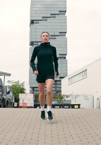 Žena v čiernom športovom oblečení behá po dlažbenéj mestkej ulici s modernou, viacpodlažnou budovou v pozadí.