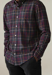 Camicia a quadri blu navy con controlli rossi e beige; presenta un colletto con bottoni, maniche lunghe e una tasca sul petto. Tessuto in cotone.