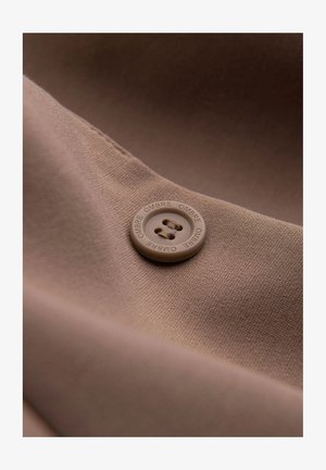 Tissu marron avec une texture lisse, présentant un bouton rond couleur tan avec le texte en relief "OMBRE" et deux petits trous pour la fixation.