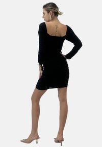 Schwarzes, gerafftes Bodycon-Kleid mit langen Ärmeln, quadratischem Ausschnitt und taillierter Silhouette. Das Model trägt beigem Sandalen mit Absatz.