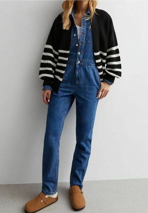 Femme portant un cardigan rayé noir et blanc sur une combinaison en denim bleu avec des chaussures marron sans lacets, debout contre un mur uni.