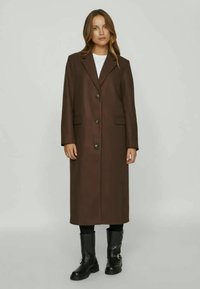 Καφέ, μακρύ παλτό από υφασμάτινο υλικό με υφή, που διαθέτει κολάρο με εγκοπή, πέντε κουμπιά και πλαϊνές τσέπες. Φορεμένο πάνω από μαύρα παντελόνια και μπότες.