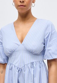 Nainen pukeutuneena vaaleansinivalkoraidalliseen mekkoon, jossa on syvä V-pääntie ja hieman puffatut lyhyet hihat, näyttäen pientä kasvojen tatuointia käsivarrellaan.