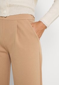 Beżowe spodnie o dopasowanym kroju z gładką fakturą, z przodu z zakładkami i bocznymi kieszeniami, zestawione z lekkim, dzianinowym kardiganem z detalami guzików.