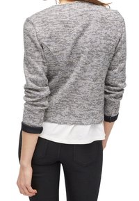 Chaqueta de manga corta, gris texturizada con un diseño corto. Presenta puños enrollados y un escote redondo, superpuesta sobre una camisa blanca.