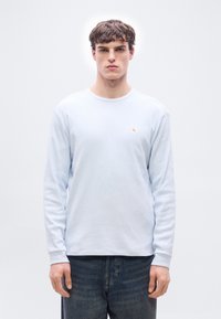  Νέος άνδρας με κοντά καστανά μαλλιά, φορούν φωτεινό μπλε πουκάμισο με μακριά μανίκια και ένα μικρό κόκκινο και λευκό λογότυπο στο στήθος, σκούρο τζιν.