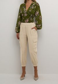 Zelená květinová blůza s nafouknutými rukávy, výstřihem do V a nařaseným pasem; krémové cargo kalhoty s bočními kapsami a zúženými manžetami; světlé béžové podpatky.