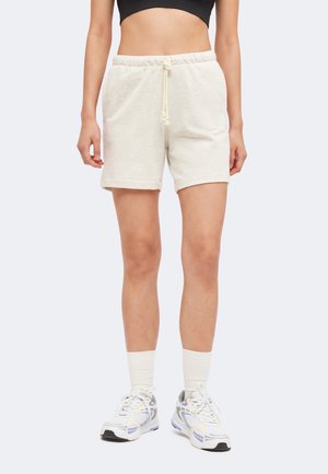 Personne portant un short beige clair à cordon, des chaussettes blanches, des baskets blanches et grises, et un top court noir, debout sur un fond uni.