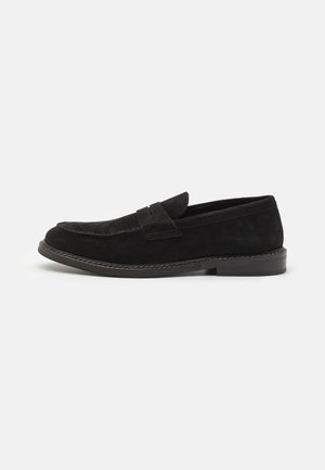 Mocassins en suede noir avec un talon plat, ornés d'une bordure cousue et d'une lanière sur le cou-de-pied. Texture lisse et design simple.