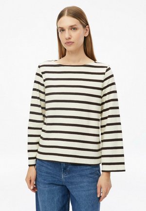 Vrouw met steil lichtbruin haar die een wit shirt met lange mouwen draagt met zwarte horizontale strepen en een blauwe spijkerbroek tegen een witte achtergrond.