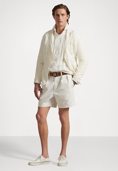 White cotton blazer over a textured white sweater, paired with beige shorts and light shoes. Features traditional stitching and casual design.