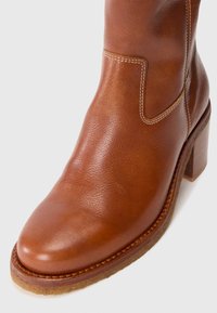 Botine din piele maro, cu vârful rotund, având un toc de înălțime medie și o cusătură albă contrastantă pe lateral.