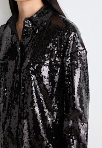 Black sequin shirt featuring a button-down collar, long sleeves, and two front pockets. The sequins create a shiny, textured surface.
