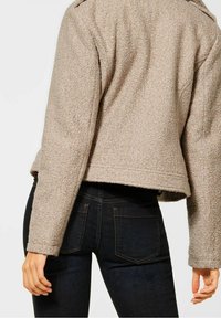 Beige texturovaná bunda s krátkým střihem a dlouhými rukávy, kombinovaná s tmavými džíny s zadními kapsami a hnědým šitím.