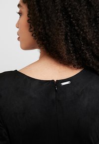 Profil d'une personne aux cheveux bouclés portant un haut noir avec une petite fermeture éclair et une étiquette métallique au niveau de l'encolure arrière.