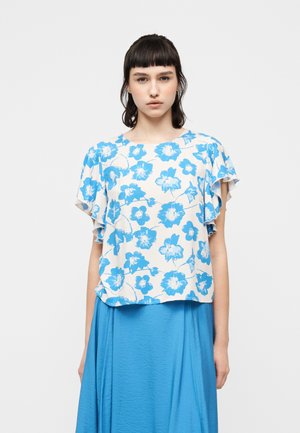 Νεαρή γυναίκα με σκούρα μαλλιά που φοράει λευκό μπλουζάκι με μπλε ανθισμένο σχέδιο και μανίκια με βολάν, συνδυασμένο με μπλε μονόχρωμη φούστα.