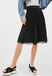 Person som bär en svart plisserad midikjol, vit topp, kort denimjacka och svarta plattformssandaler med tjocka vita sulor.