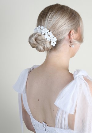 Clip per capelli floreale bianco con petali piccoli e accenti di perline, indossato in un basso chignon. Fiocchi di tessuto bianco trasparente decorano le spalle del vestito.