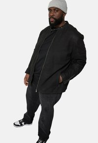 Schwarze, mit Reißverschluss versehene Jacke mit glatter Textur und Stehkragen. Trägt man über einem schwarzen Hemd und kombiniert mit abgetragenen schwarzen Jeans und Sneakern.