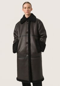 Abrigo de cuero marrón con cuello y puños de piel de oveja negra, que presenta botones, bolsillos laterales y detalles de costura horizontal.