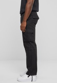 Pantalons cargo en coton noir avec une coupe droite, dotés de plusieurs poches sur les côtés et d'une texture légèrement délavée. Portés avec des baskets blanches.