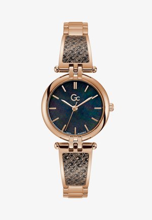 Złoty zegarek o okrągłej tarczy, ciemnej iryzującej cyferblacie i wydłużonych znacznikach godzin. Posiada metalową bransoletę z czarnym wzorem wstawki.