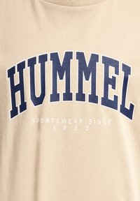 Beige bomulds T-shirt med et stort navy "HUMMEL" logo på brystet og mindre hvid tekst, der læser "SPORTSWEAR SINCE 1923" nedenfor.