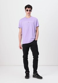 Lavendelfarbenes T-Shirt mit kurzen Ärmeln und "TOMMY JEANS"-Logo, kombiniert mit schwarzen Hosen und schwarzen Sneakers. Getragen mit runden Sonnenbrillen.