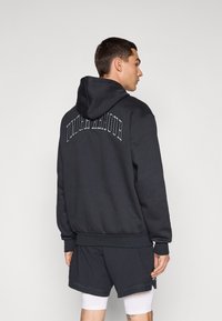 Sort hættetrøje med "UNDER ARMOUR" i hævede bogstaver på ryggen. Har en afslappet pasform og ribbede manchetter. Sammen med mørke shorts.