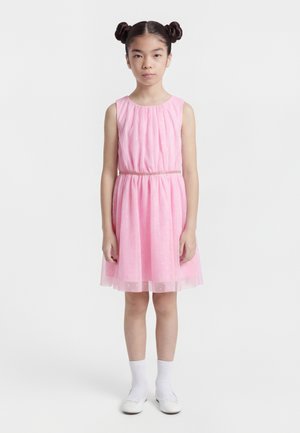 Pige med mørkt hår i to knolde iført en ærmeløs lyserød kjole, hvide sokker og hvide sko stående mod en ensfarvet hvid baggrund.