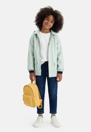 Lapsi, jolla on kiharat hiukset, pukeutuneena vaaleanvihreään takkiin, valkoiseen paitaan, sinisiin farkkuihin ja valkoisiin kenkiin, ja hän pitää keltaista reppua valkoista taustaa vasten.