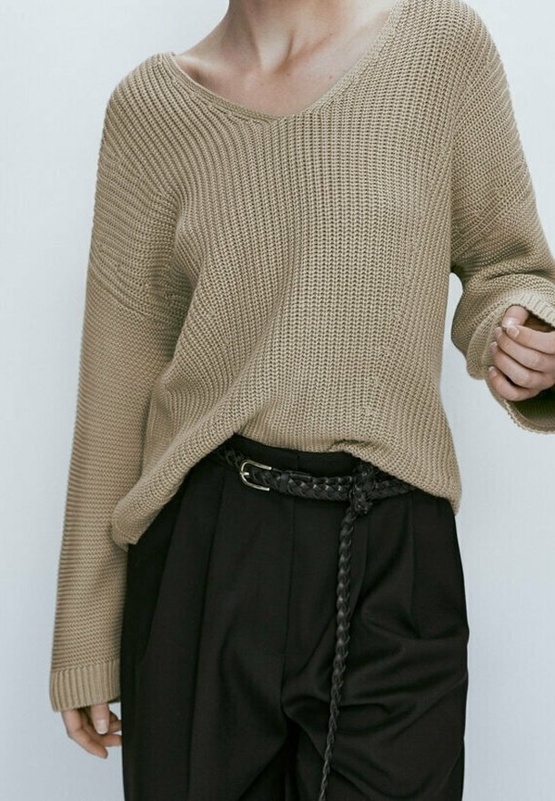 Beige knitted sweater with a loose fit, V-neck, and ribbed texture, paired with high-waisted black trousers and a braided belt.