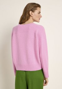 Rosa gestrickter Pullover mit entspannter Passform, geripptem Saum und Ärmeln, der eine weiche Textur aufweist. Getragen mit grünen Hosen, was einen lässigen Stil zeigt.