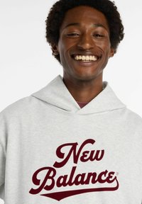 Felpa grigia con cappuccio, caratterizzata da un grande logo bordeaux "New Balance" sul davanti. Il tessuto appare morbido, con una vestibilità comoda e un collo ampio.