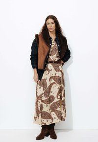 Un gilet marron et en denim sur une robe maxi à motifs paisley crème, associé à des bottes montantes marron foncé. Texture de tissu lisse, design élégant.