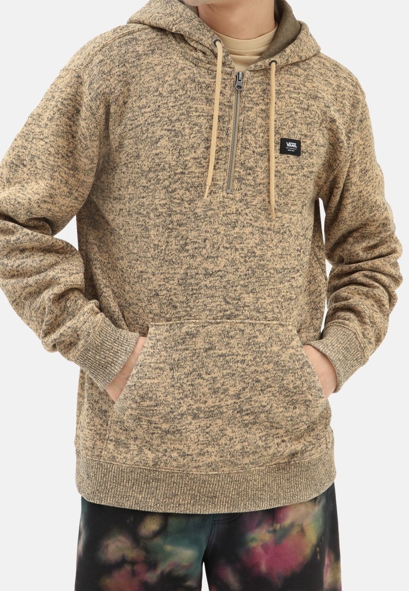 Sweat à capuche zippé beige marbré avec poche avant et cordons, porté par une personne les mains dans les poches et pantalon tie-dye visible en dessous.