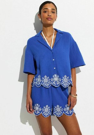 Femme portant un haut à manches courtes bleu cobalt et un short assorti avec une broderie florale blanche le long des bords festonnés.