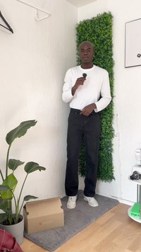 Una persona está de pie con una camisa blanca de manga larga y pantalones negros, sosteniendo un objeto pequeño. Una planta verde y una caja están cerca sobre una alfombra gris.