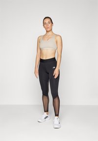 Sujetador deportivo en beige con escote redondeado y tirantes ajustables, combinado con leggings negros que presentan paneles de malla y una cinturilla ancha.