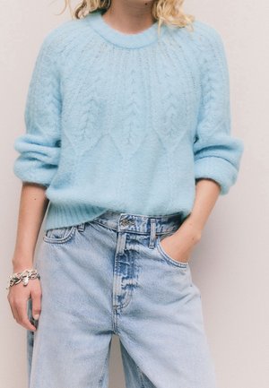 Pull en maille texturée bleu clair avec manches retroussées, associé à un jean en denim bleu clair taille haute, une main dans la poche, une personne porte un bracelet en argent.