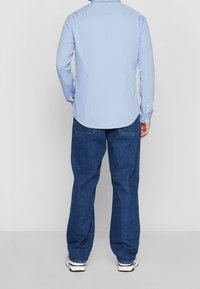 Uomo che indossa una camicia azzurro chiaro con bottoni, jeans scuri a vestibilità larga e sneakers bianche, in piedi di spalle contro uno sfondo neutro.