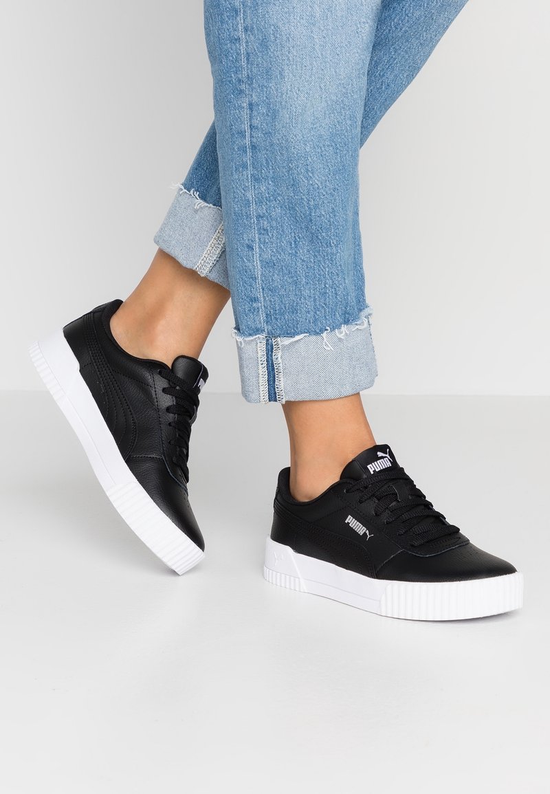 Pieds portant des baskets noires Puma avec des semelles blanches et des jeans bleu clair retroussés sur un fond gris uni.