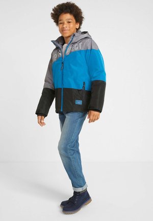 Veste colorée en bleu, gris et noir avec une capuche. Comprend une fermeture éclair, un imprimé logo et une poche latérale. Portée avec un jean bleu clair et des bottes.