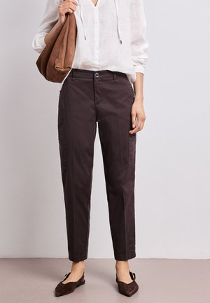 Femme portant un pantalon cargo brun foncé, un chemisier blanc, des ballerines brunes, et tenant un sac à épaule marron sur un fond uni.