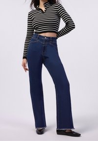 Top corto a rayas en blanco y negro con cuello abotonado, combinado con jeans de mezclilla azul de tiro alto con dobladillo deshilachado y superficie lisa.