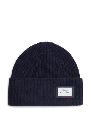 Mørkeblå strikket beanie-hue med foldet kant og et rektangulært hvidt mærke syet foran med mærkenavn.