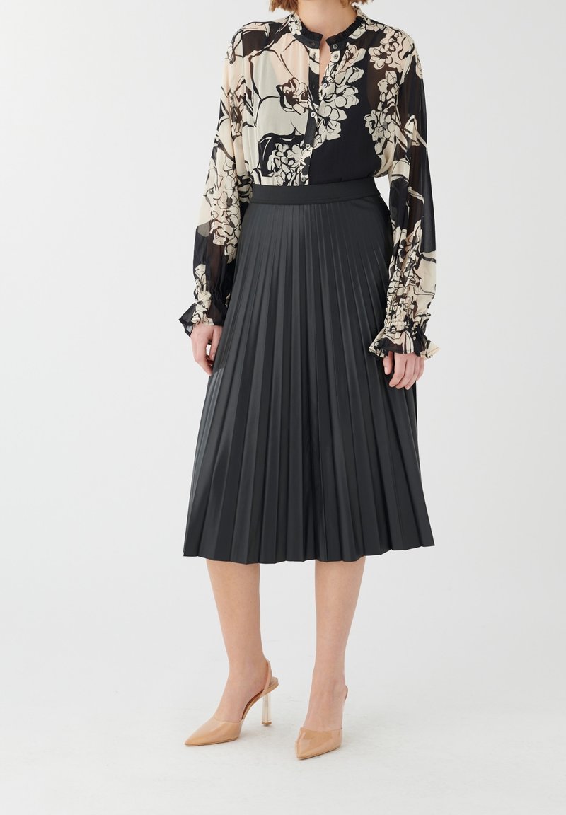 Jupe midi plissée noire associée à un blouse à motifs floraux en crème et noir, avec un tissu transparent et des manches évasées. Talons transparents en nude.