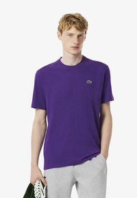 Joven con una camiseta morada de Lacoste y pantalones gris claro, sosteniendo una toalla verde y blanca con una mano, contra un fondo liso.